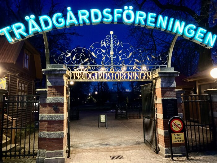 Ingång till Trädgårdsföreningen upplyst på kvällen med neontext, stängda grindar och en skylt som förbjuder cyklar i området.