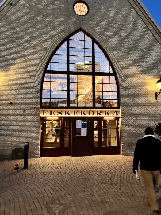 Ingången till Feskekôrka byggnad med stora fönster, tegelväggar och upplyst skylt, en person går på kullerstensväg i förgrunden.
