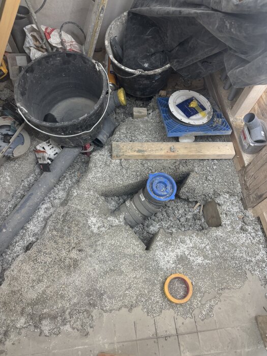 Byggarbetsplats med grå betong, blå och gul skyddande plast, och svarta hinkar. Synliga rör och fiberduk på golvet.