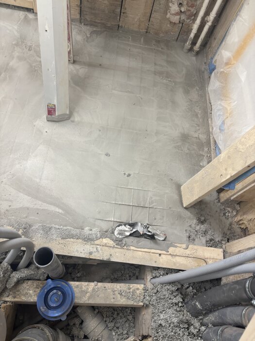 Byggarbetsplats med golv av betong eller bruk ovanför rörinstallationer, närbild på armering och plastskydd nära en vägg.