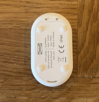 Vit, oval plastkapsling från IKEA, märkt "KLIPPBOK," med teknisk info, liggande på trägolv.