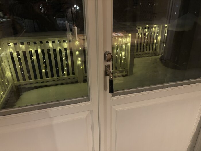 Glasdörrar sett inifrån med ett handtag på höger sida, ljusdekorationer på ett räcke syns utanför.