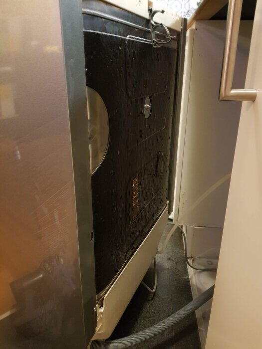 Sida av tvättmaskin med avtagbar plåt på vänster sida, sett från dess högra sida. Slang och rör är synliga bakom maskinen.