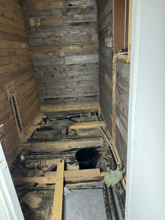 Gammalt badrum med träväggar och exponerade rör i golvet, isolerade med vadderat material, del av renoveringsprojekt.