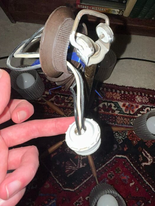 Äldre lampa med synliga elkablar och 2-poligt uttag, visas på matta med ett finger pekande på anslutningen.