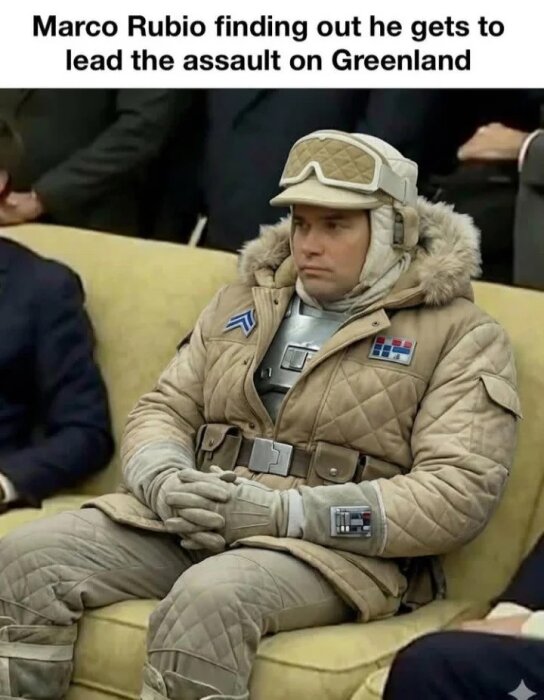 En person i en beige vinterjacka med pälskrage, sittande på en soffa i ett kontor eller konferensrum.