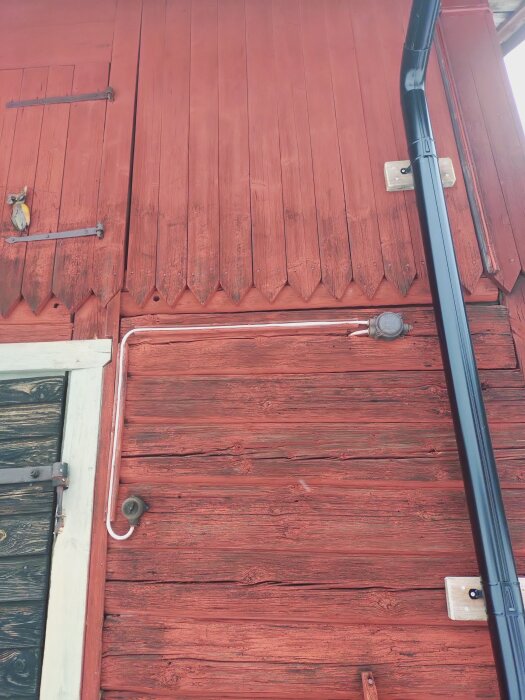 Röd träpanel med spetsiga utstående ändar på en byggnad, svart stuprör och gammalt fönsterbeslag synligt på bilden.