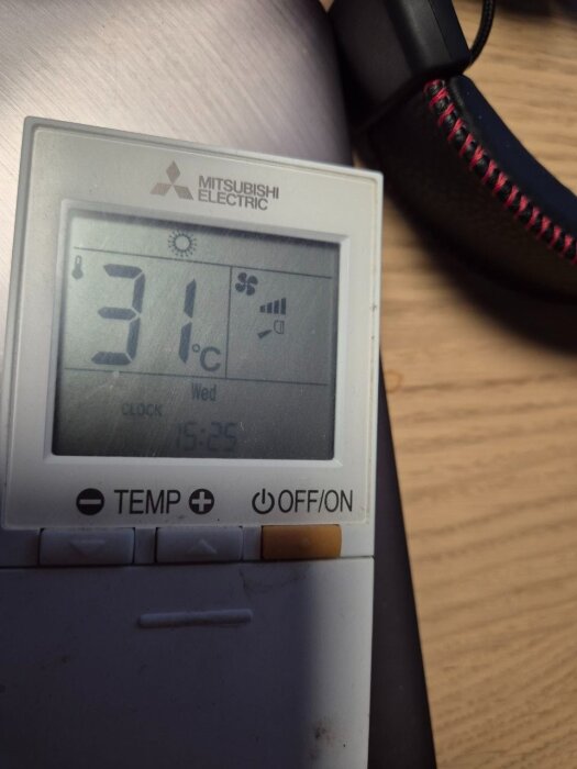 Digital termostat av märket Mitsubishi Electric visar 31 grader Celsius, med tid och temperaturinställningar, placerad på ett träbord.
