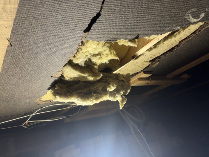 Skada på badrumsisolering där isoleringsmaterialet ligger exponerat genom ett hål i taket, med synliga kablar och trästrukturer.