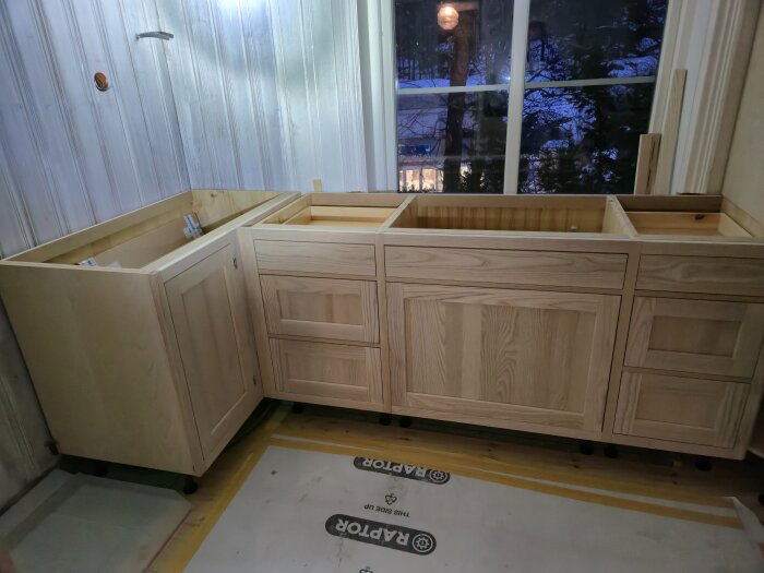Omonterade köksskåp i trä framför ett fönster under installation i ett kök under renovering.