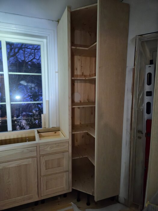 Ett kök under renovering med nya trähyllor och skåp, delvis monterade vid ett fönster.