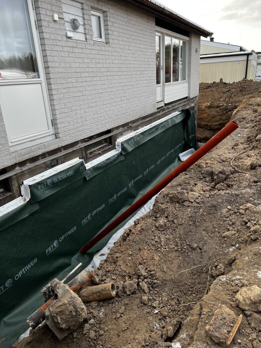 Bild föreställer en husgrund med nyligen borttaget dräneringsarbete, synliga fönster under marknivå, grönt skyddsmaterial och jordhög intill.