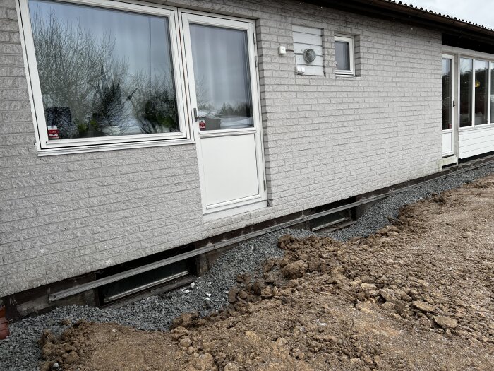 En tegelklädd källarvägg med fönster och dörr, nyligen återfylld med grus och jord efter dränering, lutande mot huset.