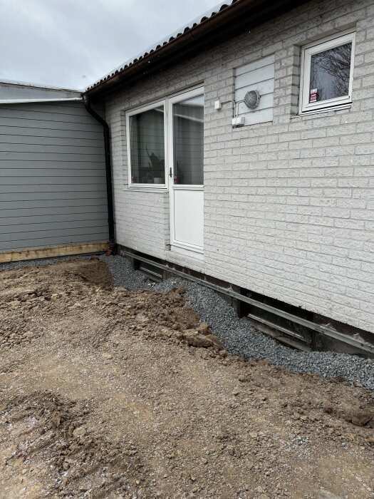 Vit fasad med källarfönster omgivna av grus och jord. Marken lutar mot huset efter dränering, och altanen är borttagen.