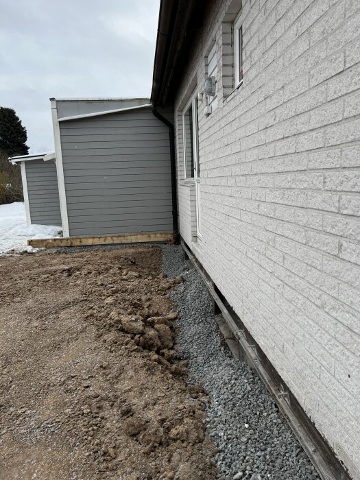 Vit tegelvägg på ett hus med gruskant och jord framför, bredvid en grå trävägg. Inledande markarbete för anläggning eller dränering.