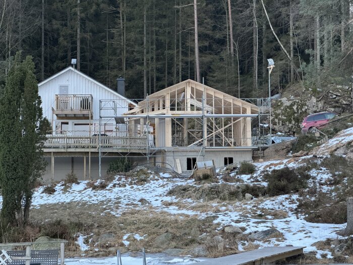 Byggarbetsplats med ofärdigt hus framför skog, snö på marken, byggställning och en bil till höger.