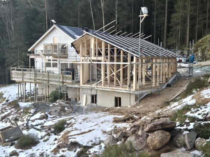 Halvbyggd trästruktur på hus bredvid skog, med byggställningar och snö på marken, under arbete för att göra det vädertätt.