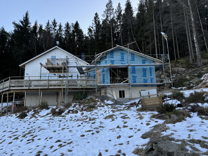 Hus under byggnation med ställningar och snö på marken, beläget vid skogsbrynet. Fönsterskydd syns på den pågående konstruktionen.