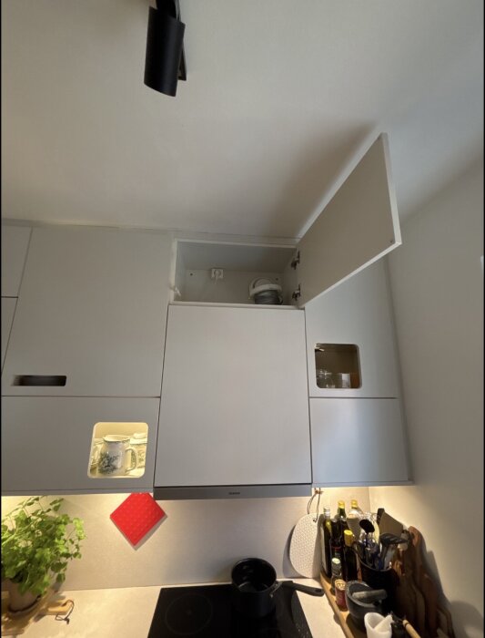 Öppet köksskåp ovanför spis med ventilationsrör inuti, bredvid porslin och hushållsartiklar. Vit köksinredning och hängande lampa syns.