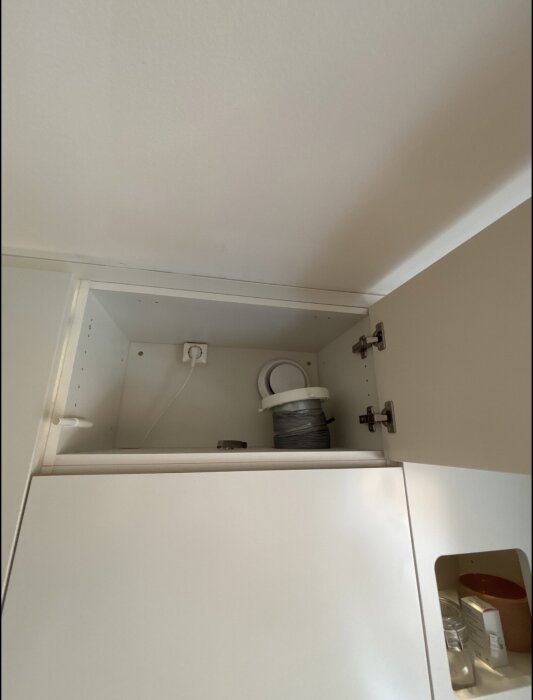 Ventilationsventil och rör inuti ett öppet köksskåp, ovanför köksfläkten, med en synlig elkontakt på väggen.