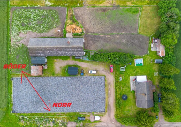 Flygfoto över gård med åkrar, en stor lada, flera byggnader och vägar. Röda pilar markerar väderstrecken norr och söder.