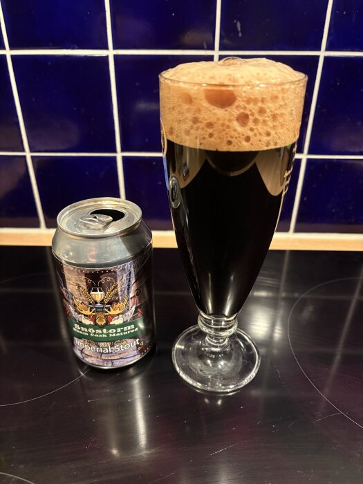 En ölburk med "Snöstorm Imperial Stout" bredvid ett glas fyllt med mörk öl och skum, mot en blå kaklad vägg.