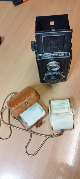 Vintage kamera med sökare, tillsammans med en brun läderväska och en vit ljusmätare, på ett bord.