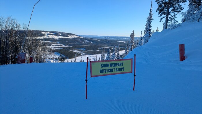 Skylt vid skidbacke markerad med "Svår nedfart" och "Difficult Slope" mot en bakgrund av snöiga träd och landskap.