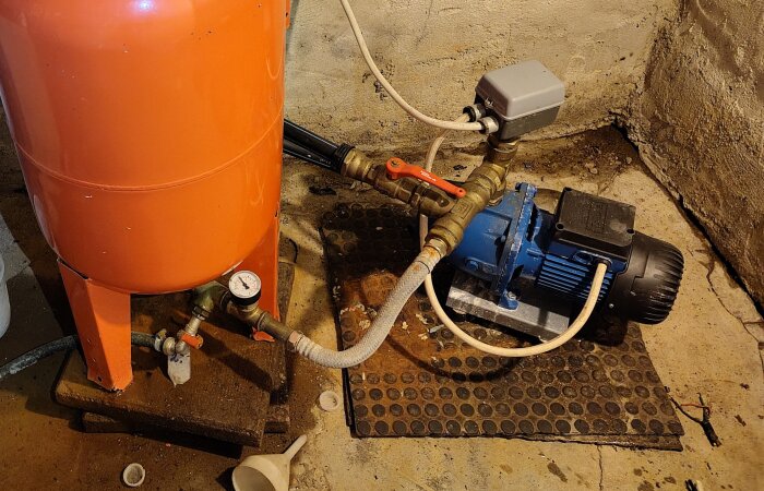 Orange hydropress och blå pump med kopplingar. Rörsystem visas med manometer och slangar på betonggolv i källarmiljö.