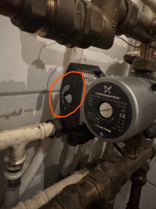 Grått reglage bredvid en cirkulationspump, markerat med orange, verkar ställt på lågt flöde i en rörinstallation.