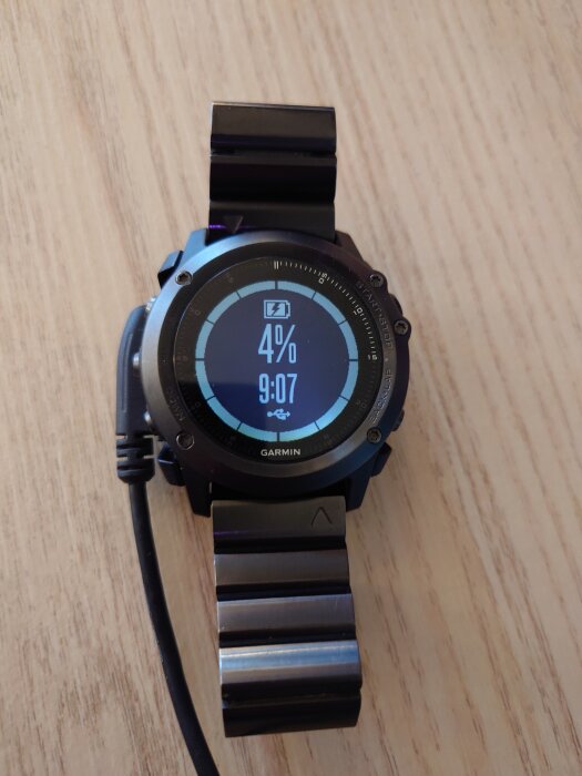 Smartklocka Garmin Fenix 3 Sapphire på trächylla med 4% batteri och laddningskabel ansluten.