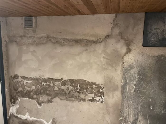 Vägg med fuktfläckar och sprickor i ett källarförråd, delvis avskavd puts med mörka partier och en ventillucka.