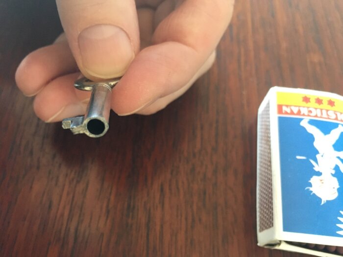 En hand håller en metallnyckel med avbruten främre tand bredvid en tändsticksask på ett träbord.