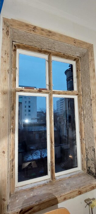 Gammalt träfönster med slitna karmar och skadad fönstersmyg under renovering i en sekelskifteslägenhet, utblick över snötäckt innergård.