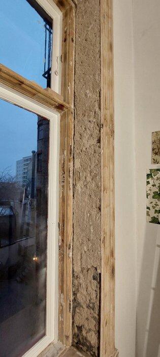 Riven fönstersmyg med avslöjad puts i en sekelskifteslägenhet, bredvid ett träfönster och stadsvy utanför.