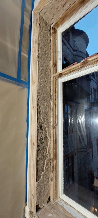 Närbild av ett fönster under renovering med exponerad träkarm och skadad puts. Bakom fönstret syns en tegelbyggnad.
