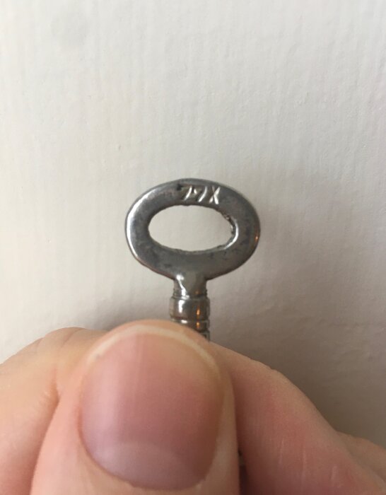 Närbild på en metallnyckel utan tänder, märkt med "77X", hållen mellan tummen och pekfingret mot en ljus bakgrund.