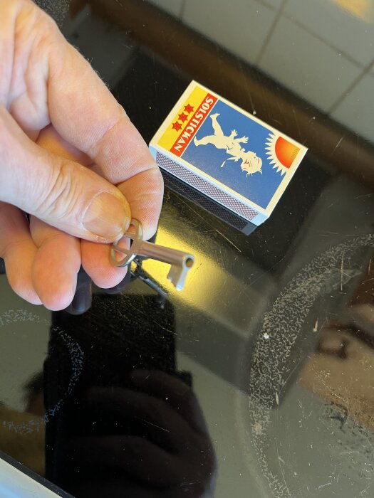 En hand håller en liten metallnyckel bredvid en tändsticksask på en mörk yta.