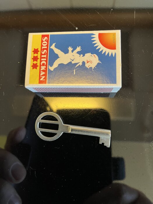 Nyckel med ovalt huvud och ett mindre centrumhål bredvid en tändsticksask märkt "Solstickan".
