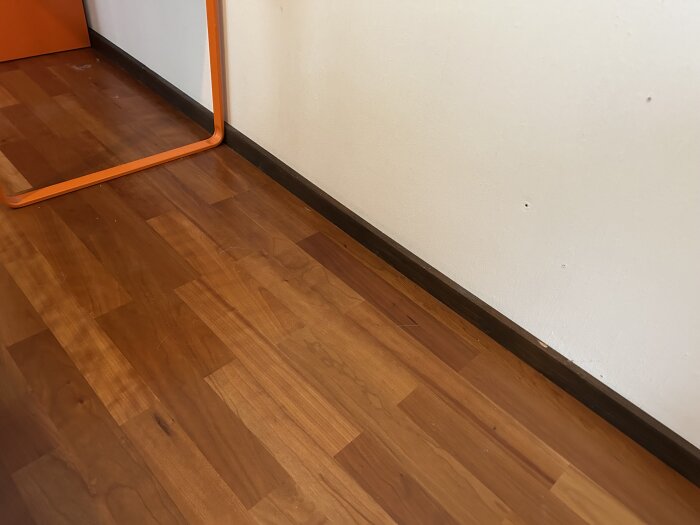 Bilden visar ett barnrumsgolv med bruna golvlister och en orange spegelram mot en vit vägg. Golvet är av trä och väggen har några små hål.