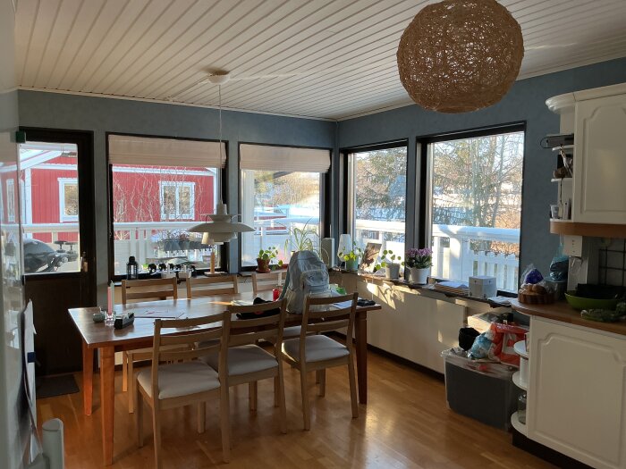 Matplats med trämöblemang, stora fönster och utsikt mot röd byggnad. Taklampa av rotting, vita väggskåp och växter i fönstret.