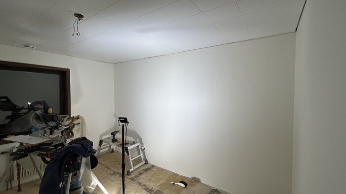 Barnrum under renovering med vita väggar och tak. Golvet är ofärdigt, och byggverktyg syns i rummet, inklusive en kap- och gersåg och stege.
