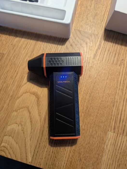 Svart och orange apparat märkt "WOLFBOX", placerad på ett trägolv bredvid en vit låda.