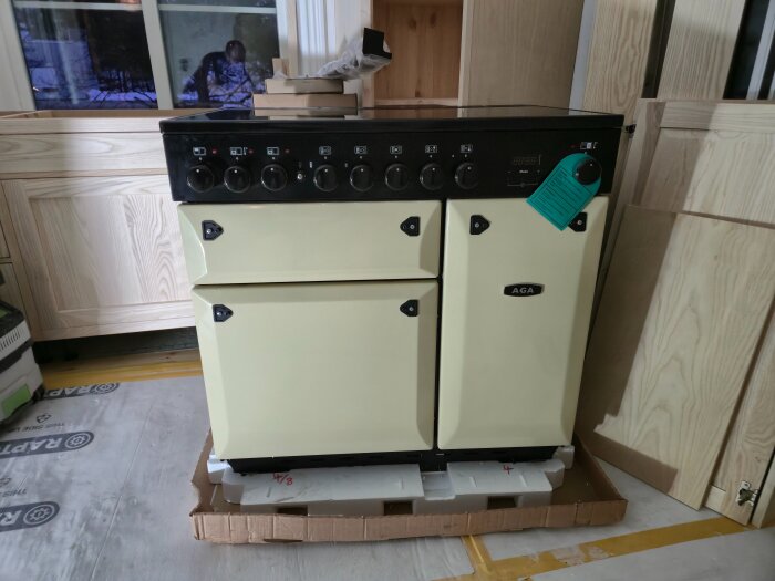 En beige AGA-spis på 160 kg installerad i ett kök under renovering, placerad på skyddspapp och kartong.
