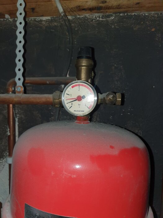Röd tryckbehållare med manometer och kopparrör mot en mörk bakgrund.