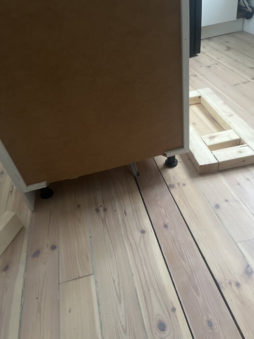 Träunderlag under köksdel, med justerbara ben synliga. Bräda på golv under köksenhet som del av stabiliseringen för inbyggnad av vinkylen.