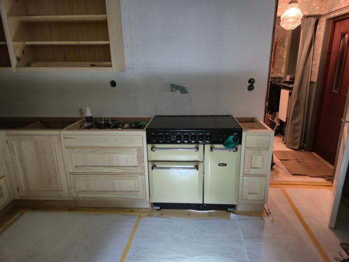Kök under renovering med träskåp, handtag och en gul Aga-spis. Vägg utan kakel och öppna hyllor ovanför.