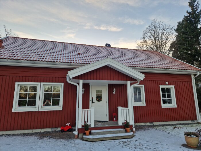 Rött trähus med vita fönster och dörr, liten veranda och snötäckt tak, planerat område för utbyggnad till höger om entrén.
