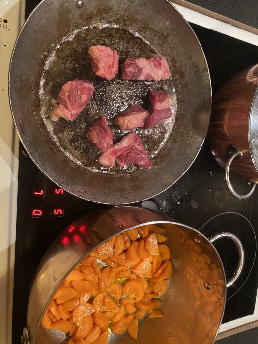 Rå köttbitar steks i panna och skivade morötter kokas i kastrull på spis, förberett för kalops.