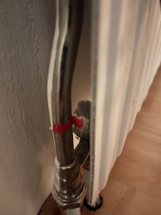 Röranslutning bakom element med rör och metallkopplingar, synlig vägg och golv, röd markering på rör.